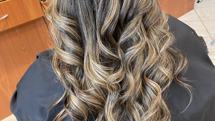 Long curls with blonde highlights over a black cape at a styling station in Queena's Haircuts