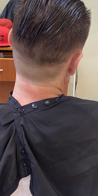 Back of a man's head with a skin fade and slicked-back top, black cape, salon station behind